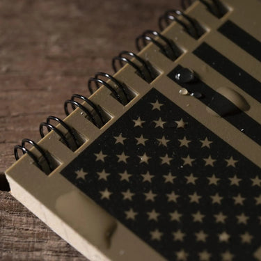Tan 3x5 Rite in the Rain top spiral notebook with black American flag design and water droplets on wood.