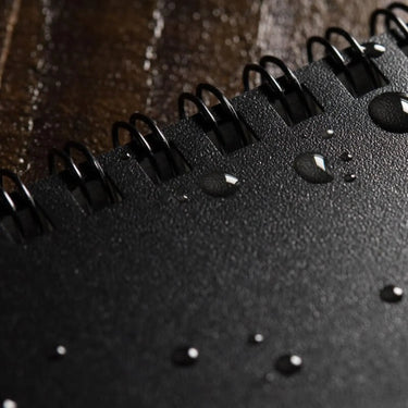 Close-up of a black, spiral-bound Rite in the Rain 3x5 notebook with water droplets on its cover.