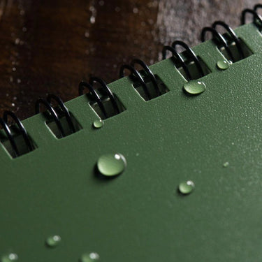 Green 4x6 Rite in the Rain top spiral notebook with water droplets beading on its durable cover.