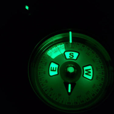Close-up of a liquid-filled lensatic compass with glowing green cardinal directions and degree markings in the dark.
