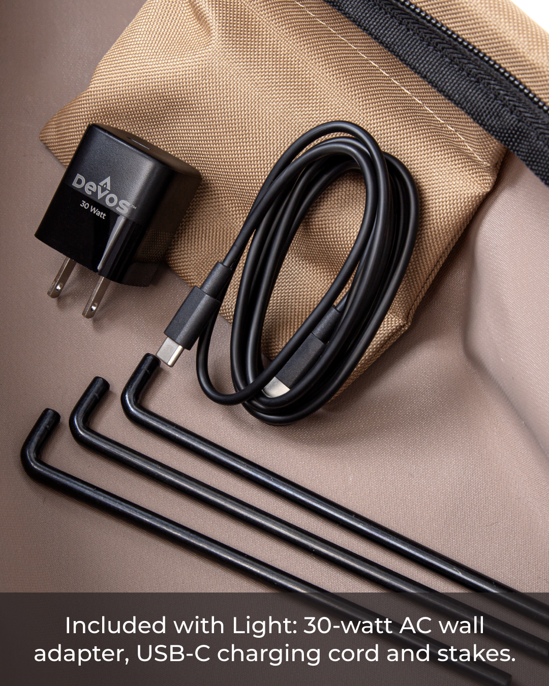 Black 30-watt Devos AC wall adapter, USB-C charging cord, and four black metal stakes on a tan fabric surface.