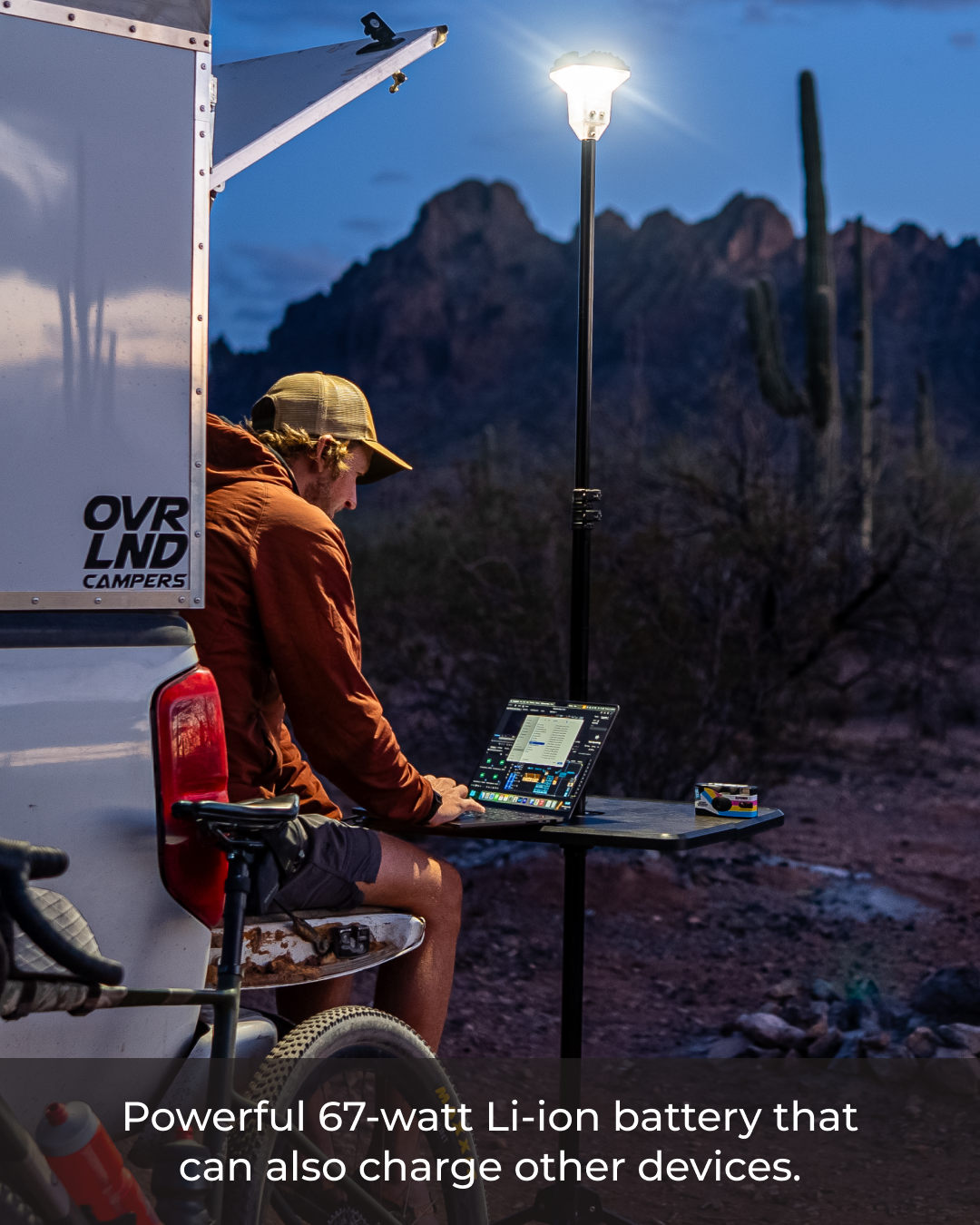 Man working on a laptop under a bright Devos LightRanger 2000 overhead light at dusk, next to a white camper.