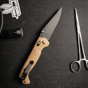 Coyote tan G10 folding knife with a black CPM MagnaCut blade and crossbar lock, shown with a flashlight and scissors.