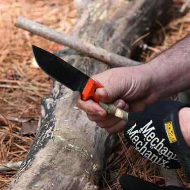 Hand holding an ESEE-4 fixed blade knife with a black blade and orange handle, carving wood outdoors.