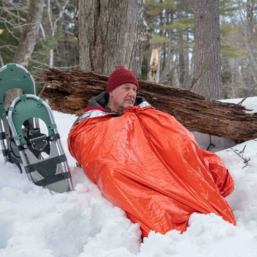 Man in a red beanie wrapped in a bright orange emergency bivvy in the snow, with snowshoes nearby.