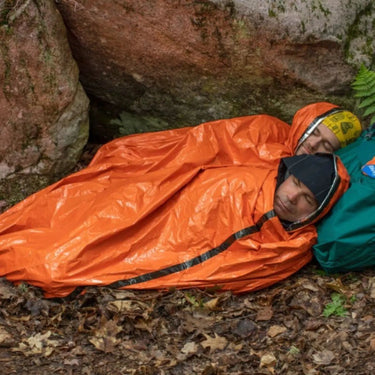 Two people sleeping in a blaze orange emergency bivvy with a rescue whistle, nestled against rocks.