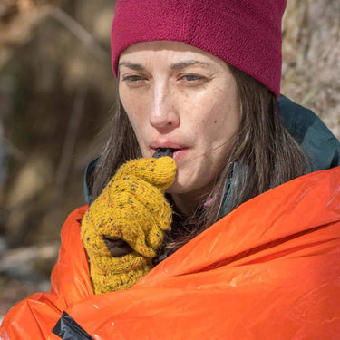 Woman in a red beanie and yellow gloves blowing a black rescue whistle while wrapped in a bright orange emergency bivvy.