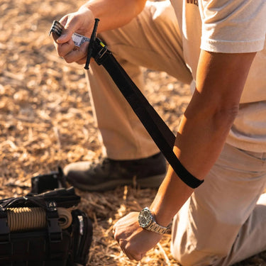 Person applying a black ETQ Gen 2 tourniquet to their arm, kneeling outdoors.