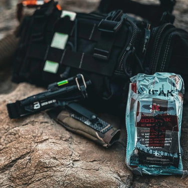 Snakestaff Systems ETQ Gen 2 tourniquet in clear packaging, next to a black tactical bag and gear on rocks.