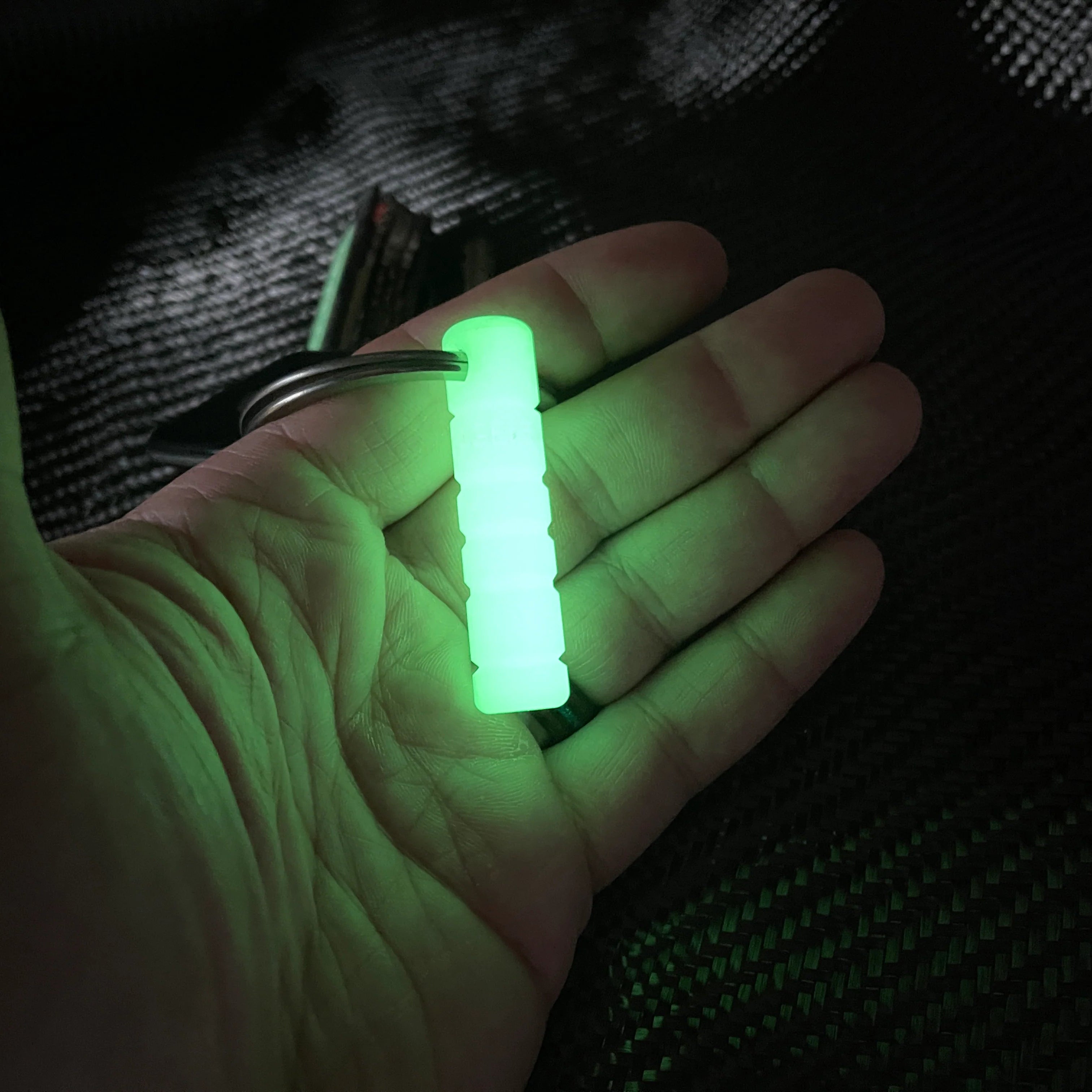 A small cylindrical keychain with a glowing green fluorescent material, held in a person's hand against a dark textured background.