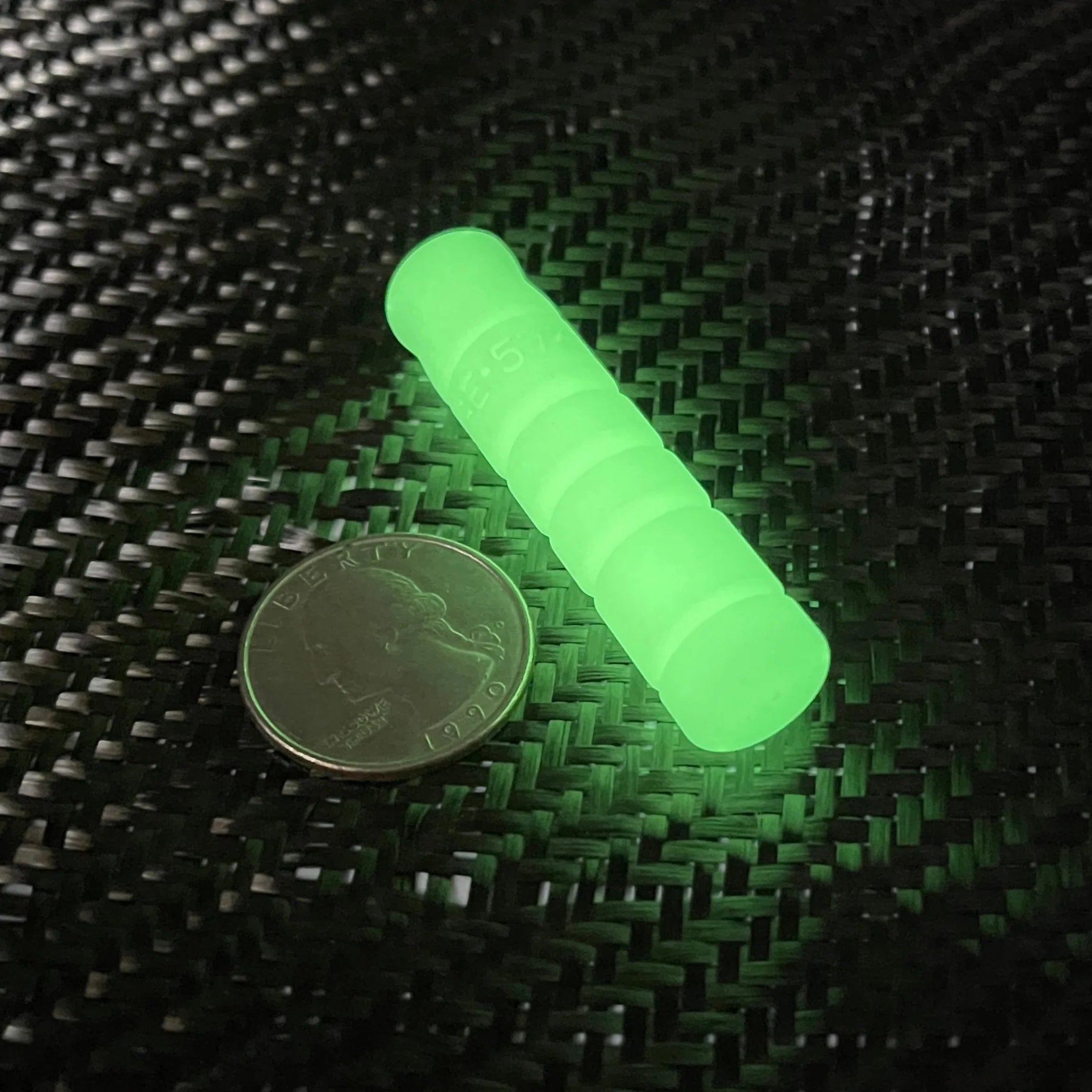 A glowing neon green cylindrical product, likely a safety beacon or light tube, shown next to a US quarter for scale on a black woven carbon fiber background.
