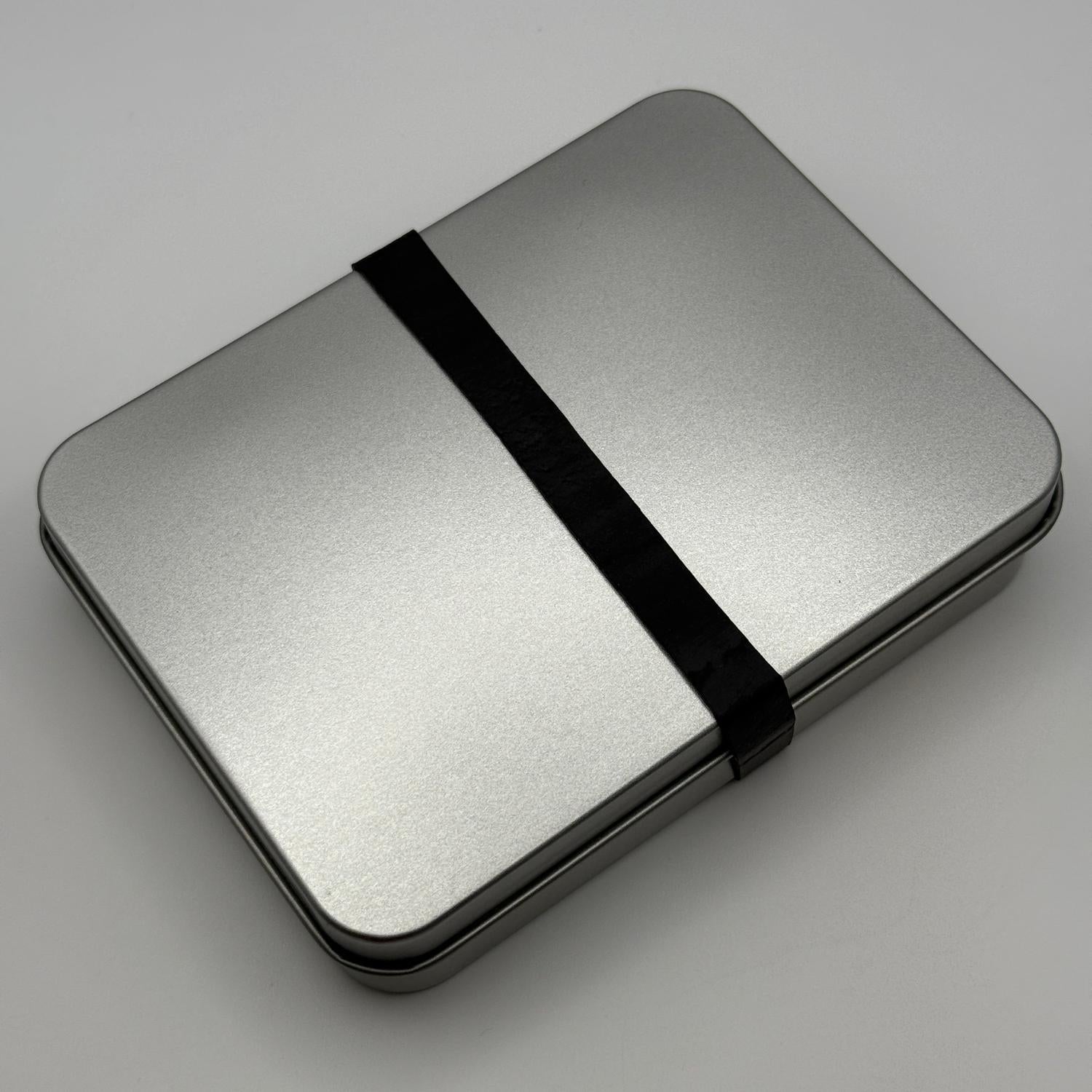 Compact, rectangular silver metal tin with a black elastic band, the INCOG emergency sustainment kit.
