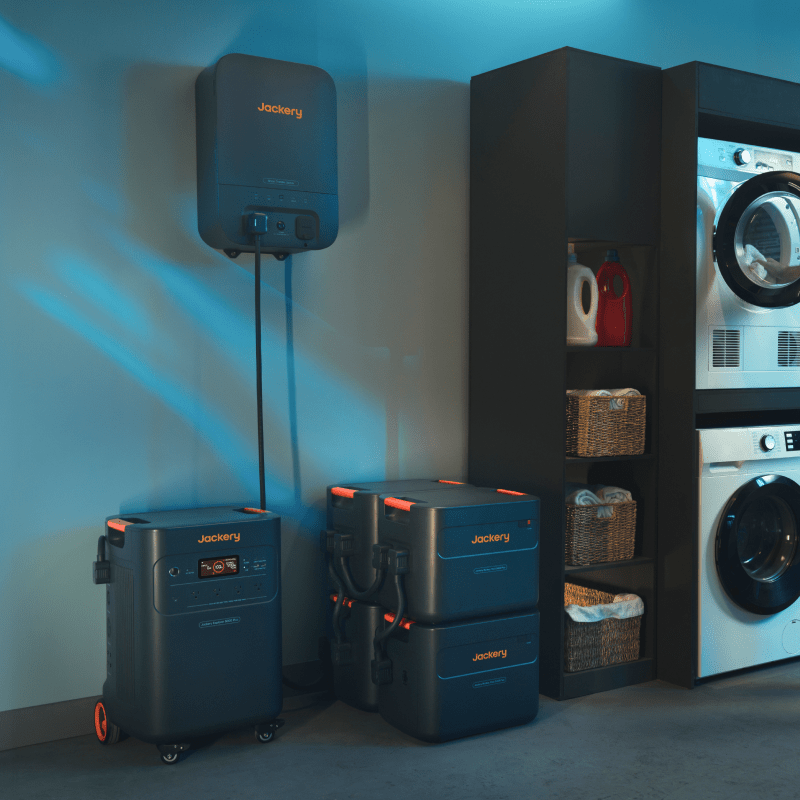 Jackery Battery Pack 5000 Plus in a dark gray finish, with orange accents, shown next to a wall-mounted unit and stacked battery packs in a laundry room.
