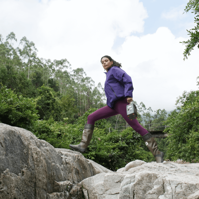 Woman in purple jacket and leggings jumping over rocks, holding a compact Jackery Explorer 300D power station.