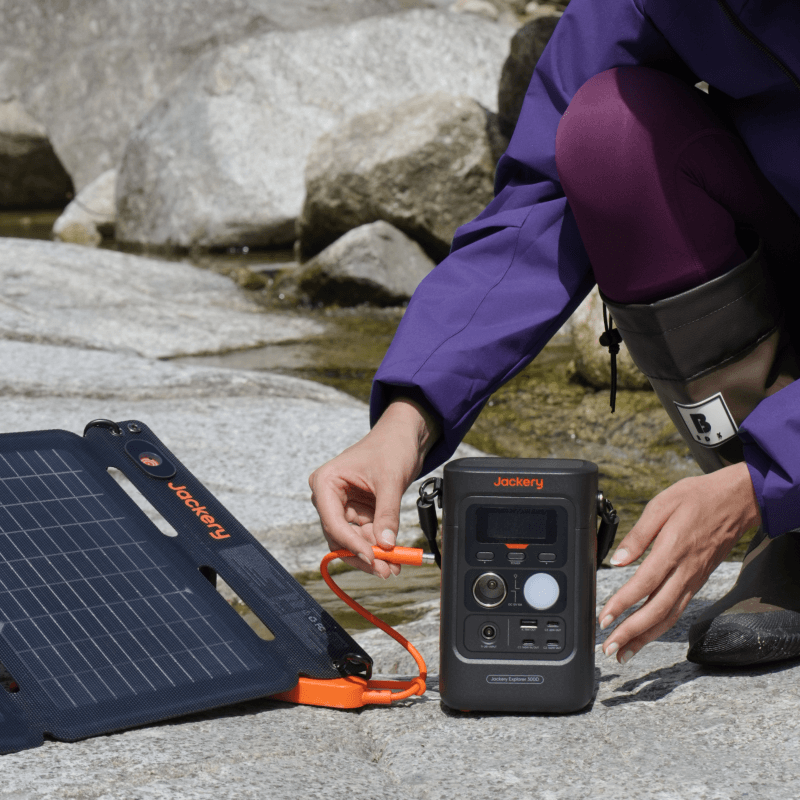 Person connecting an orange cable from a black Jackery solar panel to a compact black Jackery Explorer 300D portable power station outdoors.