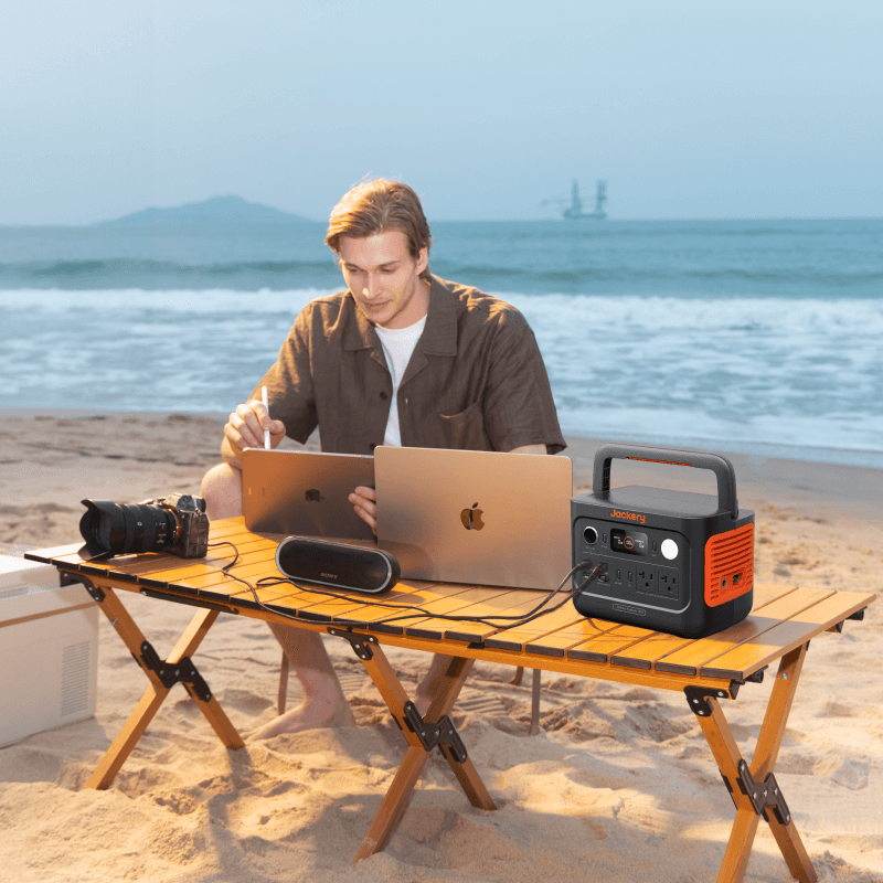 Jackery Explorer 300 v2 portable power station, black and orange, on a wooden table at the beach with a man using laptops.