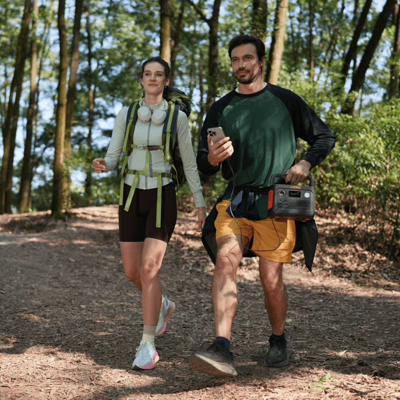 Man and woman hiking in a forest, man carrying a black Jackery Explorer 300 v2 portable power station.