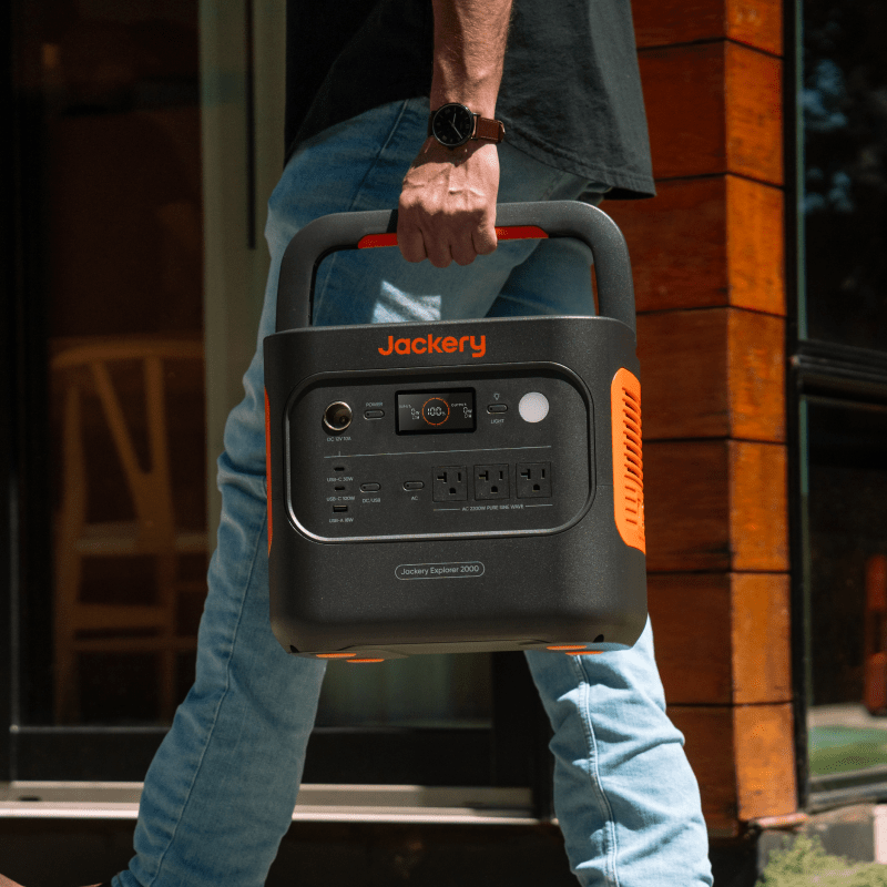 Person carrying a black and orange Jackery Solar Generator 2000 with a digital display and multiple outlets.