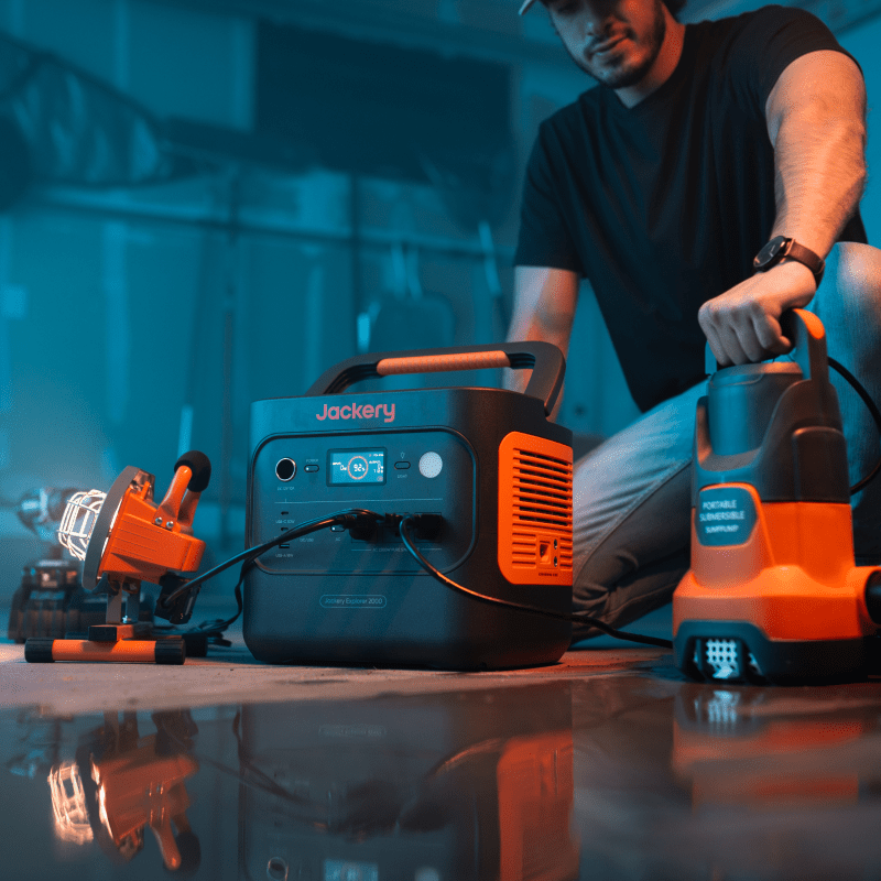 Jackery Solar Generator 2000 v2, black and orange, powering a work light and portable pump.