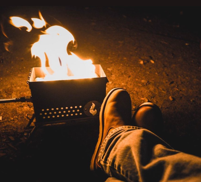 Dark metal LavaBox Campfire Kitchen with bright flames, next to a person's leg in jeans and a brown boot.