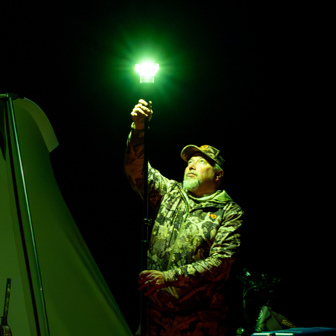 Man in camo holding a LightRanger 1200 with a bright green filter lens, illuminating a tent at night.