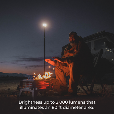 Man warming hands by a campfire at dusk, illuminated by a tall, telescoping LED area light.