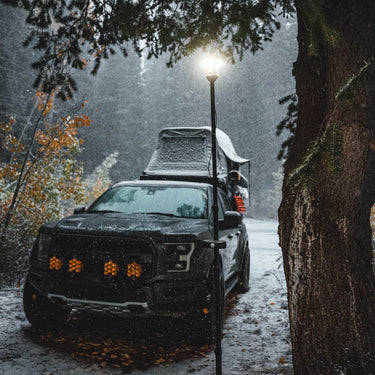 Black truck with rooftop tent and bright LightRanger pole light in a snowy forest.