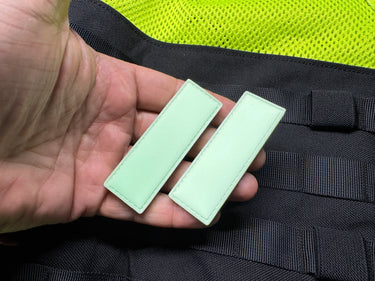 Two glow-in-the-dark, rectangular silicone Maratac Find Me Glow Tag patches held over black gear.