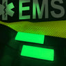 Two glowing green 1x3 inch silicone Maratac Find Me Glow Tag Patches on a dark EMS bag.