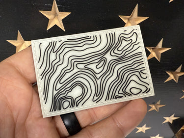 Hand holding a 3x2 inch glow-in-the-dark silicone patch with a black topographic map design.
