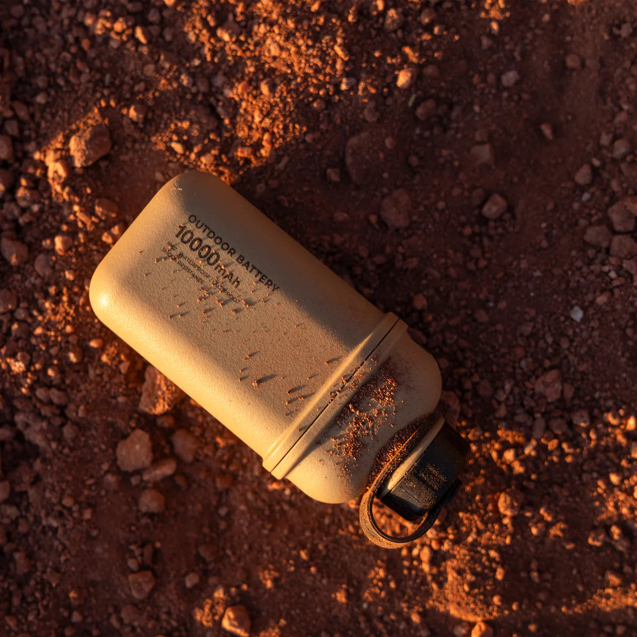 Tan NESTOUT 10,000mAh rugged outdoor power bank on reddish-brown dirt, with a black strap.