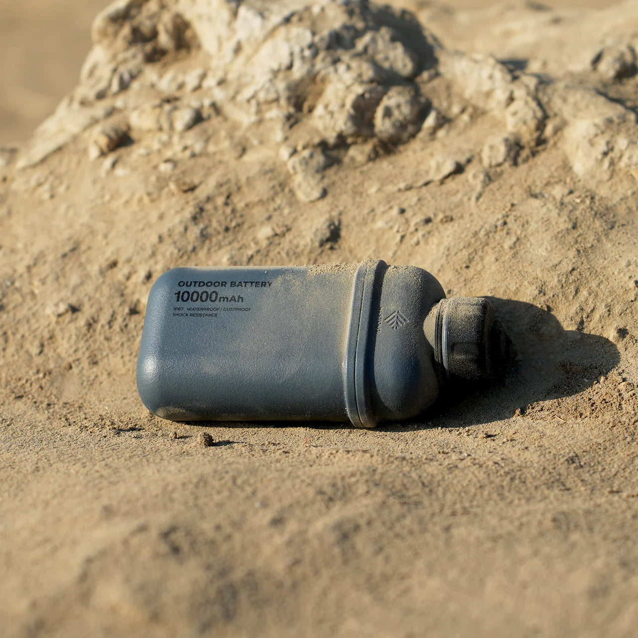 Rugged gray NESTOUT 10,000mAh power bank with a textured cap, resting on sandy ground.