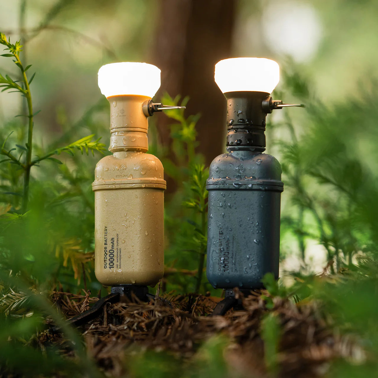 Two NESTOUT 10,000mAh power banks, one tan and one dark gray, with glowing lantern tops, standing in wet foliage.