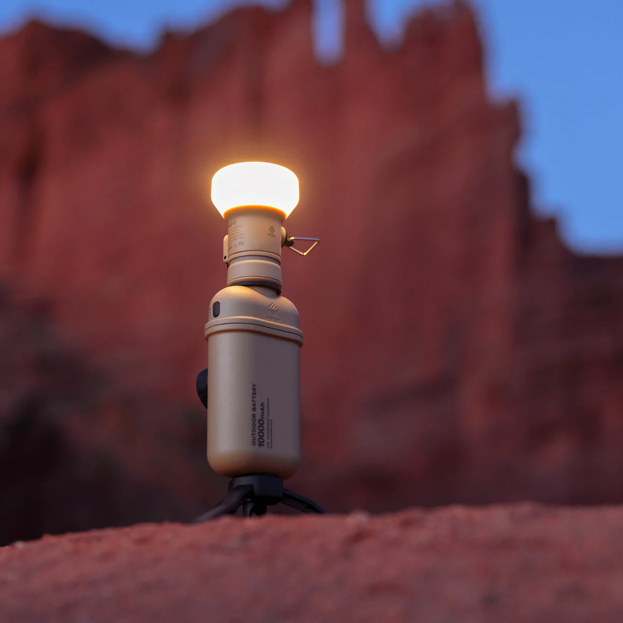 Rugged tan NESTOUT 10,000mAh power bank with an attached glowing light bulb, on a tripod against red rock formations.
