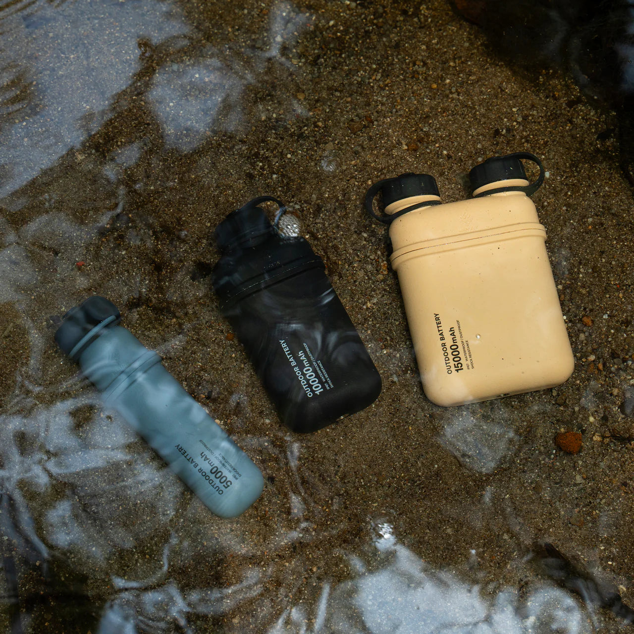 Three NESTOUT rugged power banks in black, tan, and blue, submerged in shallow water.