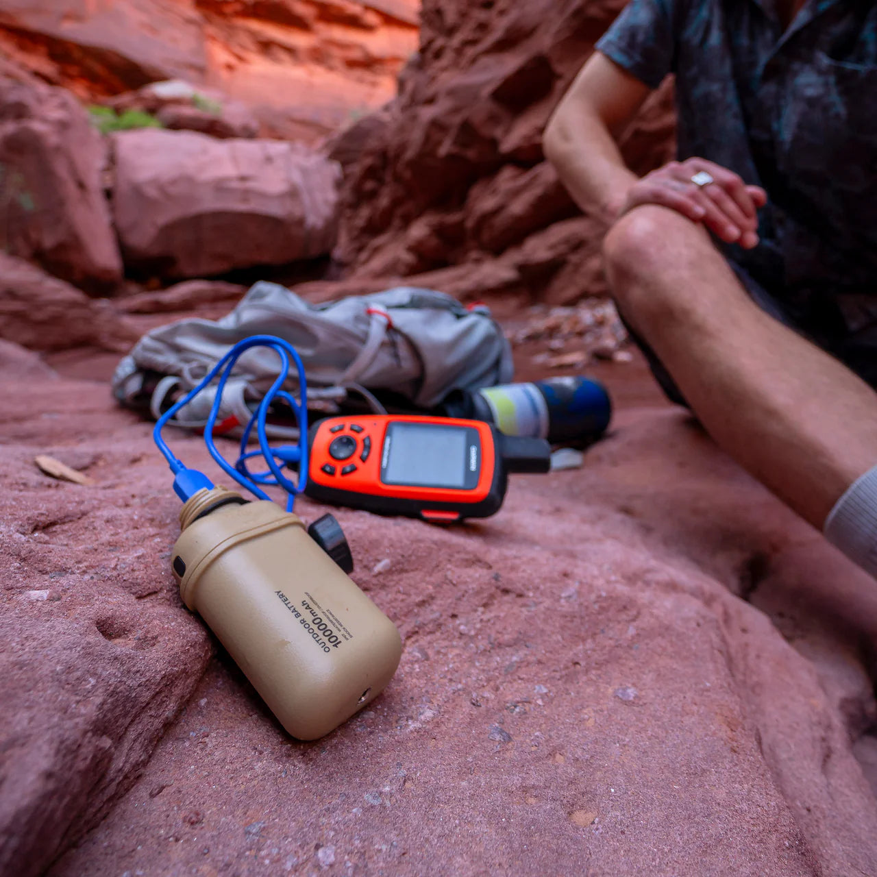Rugged tan NESTOUT 10,000mAh power bank charging a device with a blue cable on red rock outdoors.