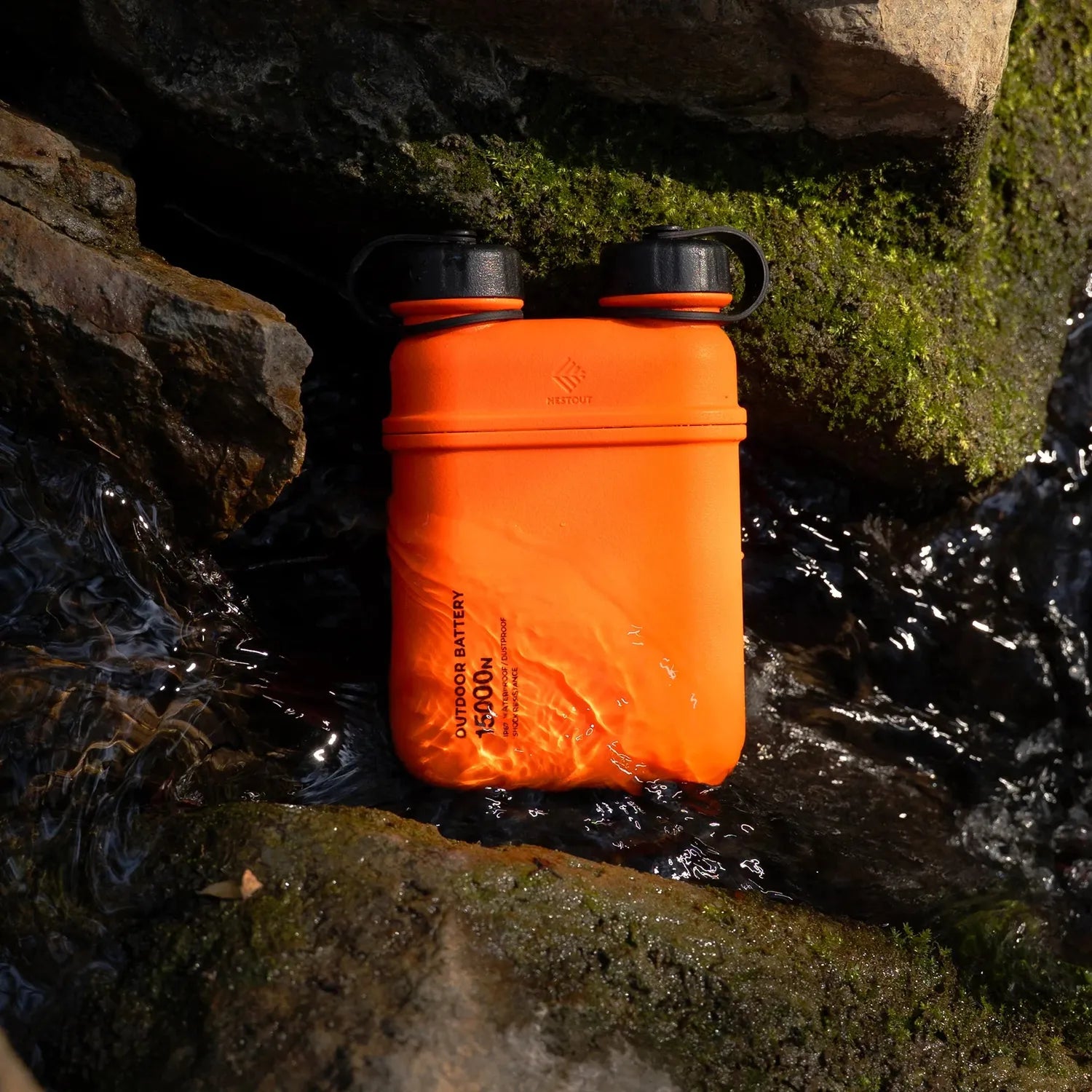 Orange NESTOUT 15000mAh outdoor power bank, water-resistant, with black caps, resting in a rocky stream.