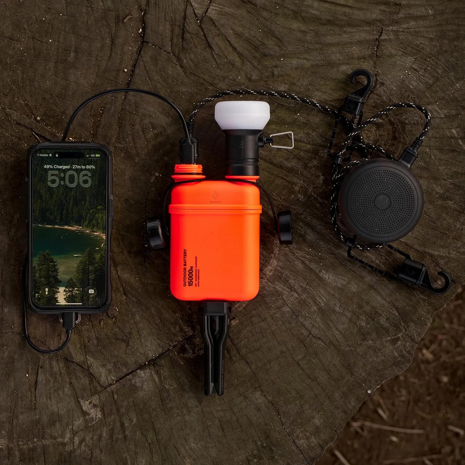 Orange NESTOUT 15000mAh outdoor power bank charging a phone, with a black speaker, on a tree stump.