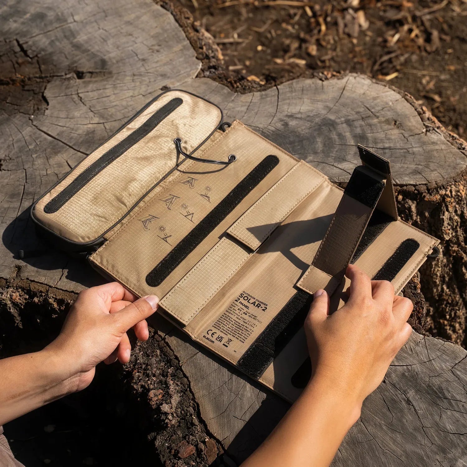 Hands unfolding a tan NESTOUT 2-Panel Solar Charger V2 on a tree stump, showing its compact, portable design.