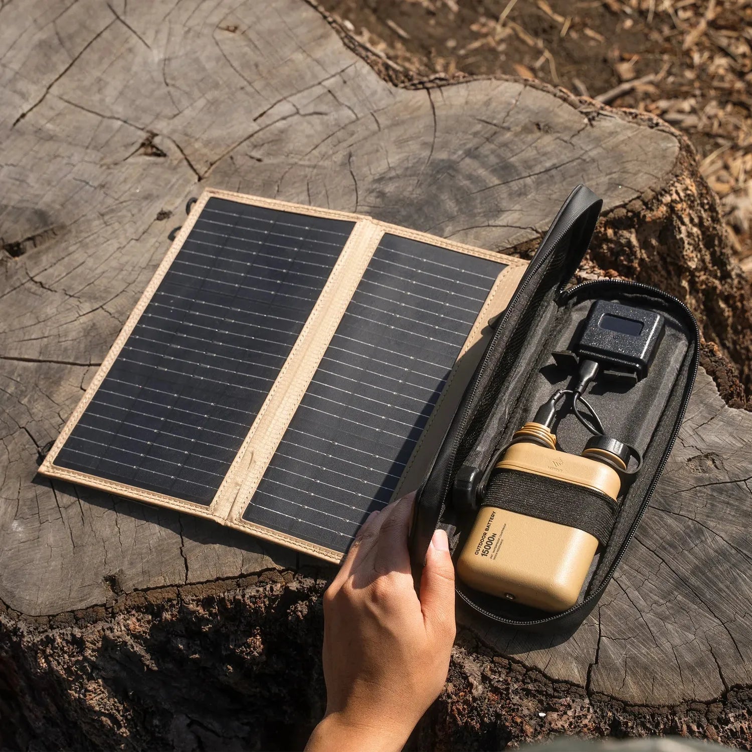 NESTOUT 2-Panel Solar Charger V2, tan and black, open on a tree stump next to a hand holding a tan battery pack.