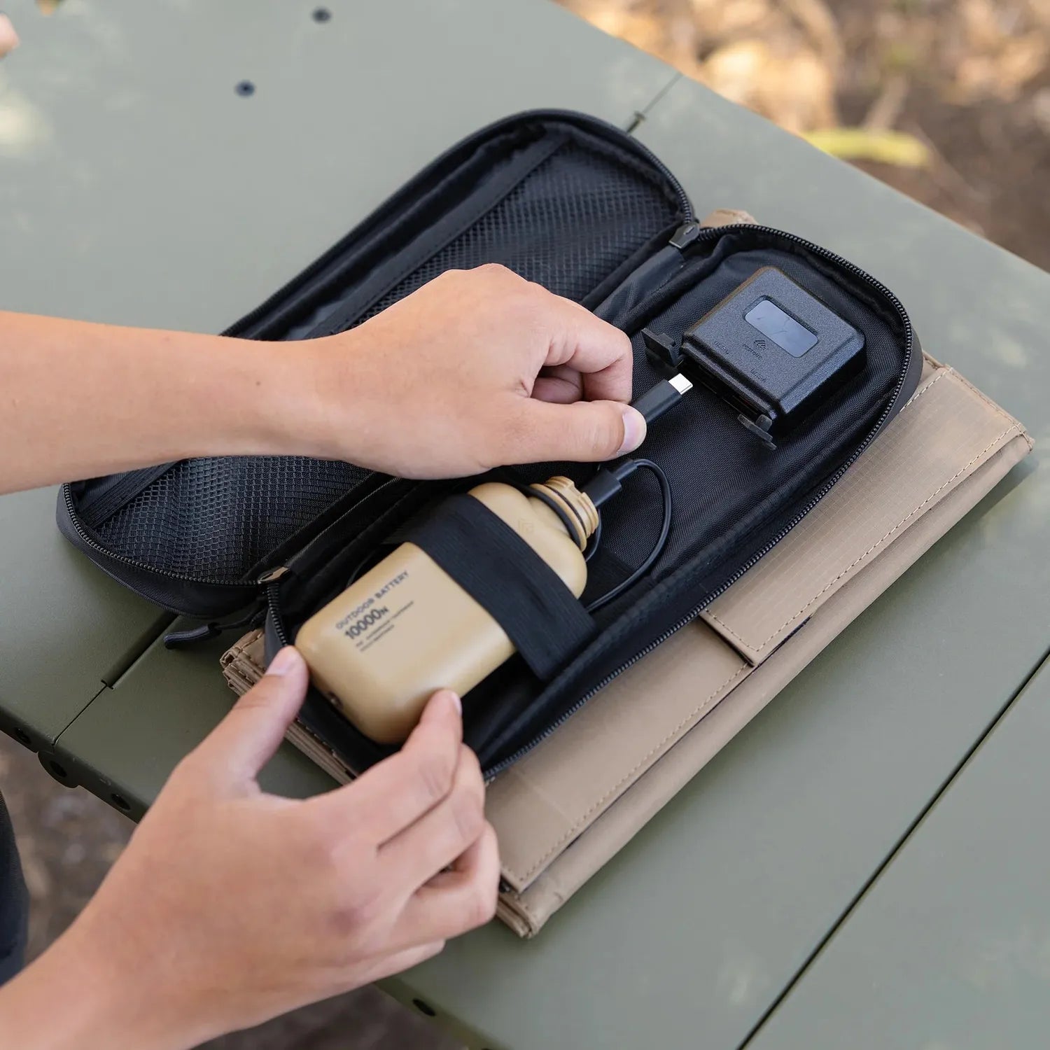 Hands connecting a black USB-C cable to a black device inside a NESTOUT 4-panel solar charger's black zippered pouch, next to a tan battery.