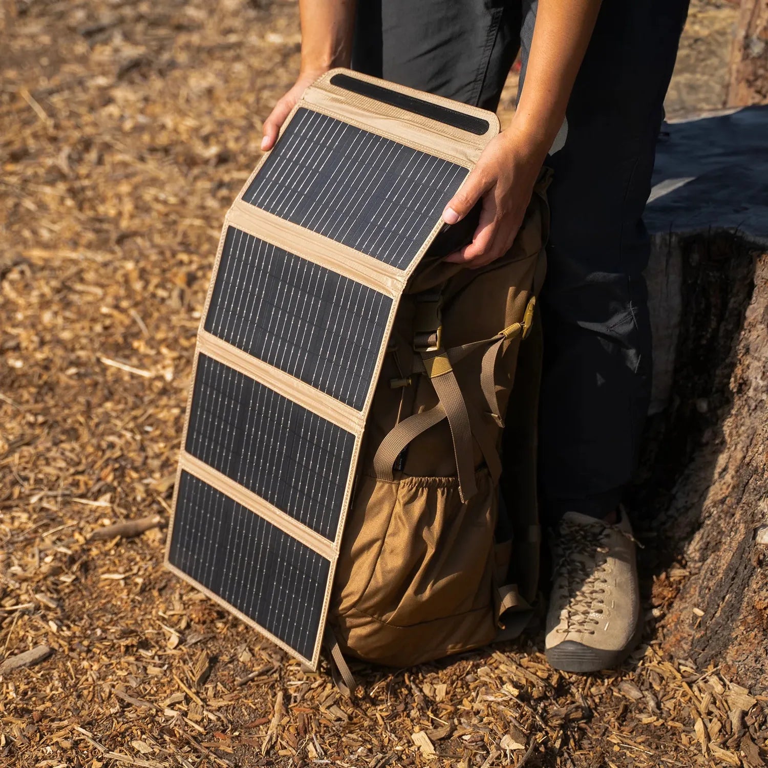 Person holding a folded NESTOUT 4 Panel Solar Charger V2 with black monocrystalline cells and tan fabric, next to a backpack outdoors.