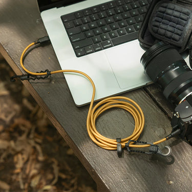 Tan braided USB-C cable with black connectors and hooks, coiled on a wooden table next to a laptop and camera.