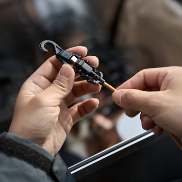 Hands hold a tan braided USB-C cable with a black rugged connector and protective cap with a hook.