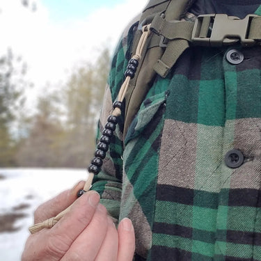 Hand holding black pace count beads on tan cord, attached to a green plaid shirt and tactical backpack.