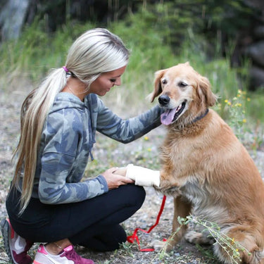 Blonde woman in camo hoodie tending to a golden retriever's bandaged paw outdoors.