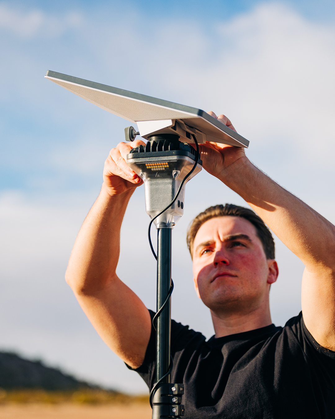 Man adjusting a gray Starlink Mini dish mounted on a black LightRanger pole under a blue sky.